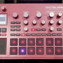 Červený sampler KORG electribe sampler MUSIC PRODUCTION STATION. Zařízení obsahuje displej, několik otočných knoflíků a mřížku podsvícených tlačítek.…