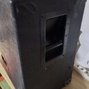Černý reproduktorový box s povrchem z texturovaného materiálu. Na hranách jsou umístěny plastové ochranné rohy a na boku je integrovaná rukojeť v pla…