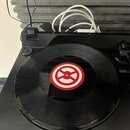 Černý gramofon s umístěnou černou vinylovou deskou. Uprostřed desky je červenobílý kulatý štítek s grafickým symbolem. V pozadí jsou vidět bílé a čer…