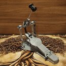Stříbrný kovový pedál basového bubnu s řetízkovým pohonem a černým plastovým knoflíkem nahoře. Na bocích stupně je vyryt nápis RONVAN. Povrch kovu je…