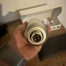Stříbrný kovový válečkový objekt s konektorem v čele. Na boku je vidět část nápisu "POD". V pozadí se nachází bílé papírové krabičky, na jedné z nich…