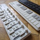 Tři MIDI kontroléry v bílé a černé barvě položené na dřevěné podlaze. Nejlevější bílý kontrolér v popředí obsahuje posuvníky, knoflíky a tlačítka s o…