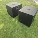 Dvě černé basové bedny s texturovaným povrchem umístěné na trávě. Každý box má na horní straně kulatý otvor pro instalaci tyče a na boku integrovanou…