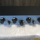 Modrý rackový procesor s černým horním krytem a řadou ovládacích knoflíků. V levém horním rohu je červené logo "Warm Audio" a nápis "TUBE EQ". Na pan…