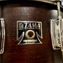 Detail tmavě hnědého dřevěného korpusu bubnu s chromovaným kováním. Uprostřed je černý štítek s bílými nápisy TAMA, SUPERSTAR ® a MADE IN JAPAN. Pod…