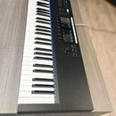 Digitální piano v šedém dřevěném korpusu s černým ovládacím panelem. Nástroj má standardní klávesnici s bílými a černými klávesami. Na ovládacím pane…