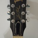 Černá hlava elektrické kytary s nápisy v žluté barvě. V horní části je logo Gibson a pod ním nápis Les Paul JUNIOR. Nástroj má šest chromovaných ladi…