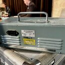 Šedý kovový přístroj s rukojetí na horní straně a ventilačními mřížkami vzadu. Na zadní straně je napájecí konektor a dvě nálepky s nápisy VEB Musike…
