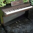 Digitální piano v tmavě hnědém dřevěném provedení s bílými a černými klávesami. Na horním楽谱stavu je vyveden nápis CELVIANO. Nástroj stojí venku na zp…