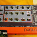 Červený syntetizátor s nápisem nord rack virtual analog. Na předním panelu jsou rozděleny sekce OSC 1, OSC 2, AMPLIFIER a FILTER s řadou otočných kno…