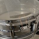 Detailní pohled na chromovaný snare drum s bílým koženem. Na bubnu je umístěna kovová mřížka (snare mute) a k okraji je připevněn držák s nápisem "Pe…