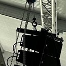 Fotografie zobrazuje lineární pole pěti černých reproduktorů zavěšených na kovovém řetězu z modrého zdvigače. Celý systém je upevněn k hliníkovému tr…