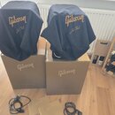 Dvě černé textilní krycí plachty s nápisy „Gibson“ a „Les Paul“. Plachty jsou položeny na dvou hnědých papírových krabicích, z nichž každá má uprostř…