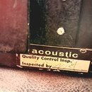 Detailní záběr na dvě nalepené štítky na tmavém povrchu. Horní černý štítek nese bílý nápis acoustic, spodní bílý štítek obsahuje text Quality Contro…