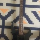 Černá elektrická kytara s tmavým hnedým hmatníkem. Na hlavě je viditelné zlaté logo Banex. Nástroj má dva humbuckery a tremolo mechanismus. Povrch ky…