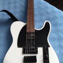 Bílá elektrická kytara s černým pickguardem a hnědým hmatníkem. Nástroj je vybaven dvěma snímači typu humbucker a černým mostem. Na hlavě krku je vid…