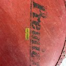 Detail červeného povrchu bicího nástroje s černým nápisem Premier. Uprostřed je nalepený žlutý štítek s textem MUSIKHAUS FRANZ GOTRUBA WIEN 12 EICHEN…