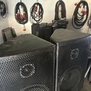 Dva černé reproduktory s kovovými perforovanými mřížkami. Povrch skříně je texturovaný a v horní části jsou vidět prach a drobné nečistoty. Na mřížká…