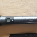 Šedý bezdrátový mikrofon s černou mřížkou. Na těle je bílý nápis SENNHEISER a pod ním XS WIRELESS. V horní části je malý displej, tlačítko MUTE a čer…