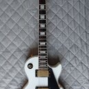 Bílá elektrická kytara s černým hmatníkem a černým pickguardem. Na hlavě je bílý nápis Epiphone. Nástroj má zlaté potenciometry, ladící mechanismy a…