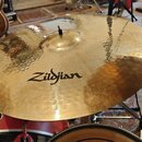 Zlatavý kovový činel s viditelným kovánívým povrchem a mírným opotřebením. Uprostřed je černý nápis Zildjian. Činel je osazen na stojanu v rámci bicí…