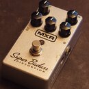 Stříbrný efekt pedál s pěti černými oválnými knoflíky. Na horní straně je logo MXR a nápis Super Badass DISTORTION. Jsou zde viditelné popisky OUT, I…