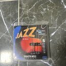Balení basových strun v průhledném plastovém obalu. Na obalu je nápis JAZZ Electric Bass, Flat Wound a značka THOMASTIK-INFELD VIENNA. Je zde uvedena…