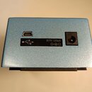 Modrý kovový box s dvěma konektory na zadní straně. Je zde USB port a vstup pro napájení s nápisem DC9V 500mA a symbolem polarity. Povrch je jemně te…