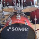 Červená bicí souprava s kovovým hardwarem. Na přední koži basového bubnu je bílé logo a nápis SONOR®. Na držáku tomů je viditelný nápis SONOR. Povrch…