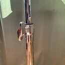 Detail chromovaného kovového stojanu s mechanismem pro regulaci výšky. Na spojovací části je viditelný utahovací šroub a černý gumový kroužek. Povrch…