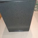 Černý subwoofer s kovovou perforovanou mřížkou přes celý přední panel. V dolní části je umístěn oválný štítek s nápisem tce a označením modelu SUB 18…
