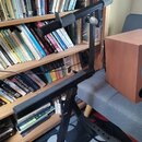 Černý kovový stojan na noty s teleskopickým ramenem a horní opěrnou lištou. Stojan je umístěn v interiéru vedle knihovny a šedého pohovku. Povrch lak…