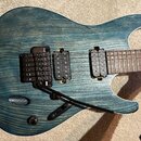 Elektrická kytara s modrozeleným povrchem s viditnou strukturou dřeva a bílým lemováním. Nástroj má dva černý humbuckery, černý most s pákou a jeden…
