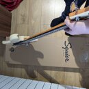 Bílá elektrická basová kytara s hnedým hmatníkem a světlou hlavou. Na kartonové krabici pod nástrojem je nápis Squier BY FENDER. Kytara je držena ruk…