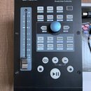 Černý kontrolér PreSonus FADERPORT Production Controller. Zařízení obsahuje posuvník s označením dB a U, modrý otočný knoflík a sadu bílých tlačítek…