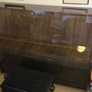 Hnědé pianino s vysokým leskem a dřevěnou strukturou. Před nástrojem stojí černá pianistická židlička s čalouněným sedadlem. Na nástroji ani v jeho o…