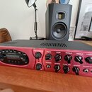 Červený kytarový zesilovač s černým horním krytem. Na předním panelu jsou nápisy POWER, AMP MODELS, DRIVE, BASS, MIDDLE, TREBLE a PRESENCE. V levé čá…