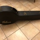 Černý tvrdý kufýr na hudební nástroj s kovými zapínacími zámky a rukojetí. Na přední straně je umístěn zlatý nápis Gibson. Povrch kufýru vykazuje mír…