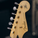 Záběr na hlavu kytary z přírodního světlého dřeva se zlatými ladičkami. Na hlavě je čitelné logo Fender a nápis STRATOCASTER s doplňkem "WITH SYNCHRO…