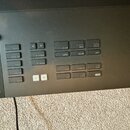 Detailní záběr na ovládací panel černého elektronického hudebního zařízení. Jsou vidět řady šedých tlačítek s bílými nápisy, mezi nimiž jsou napříkla…