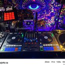 Fotografie zachycuje DJ set umístěný před barevným gobelínem s motivem oka. Uprostřed je černý DJ kontrolér se značkami RANE a serato, vybavený dvěma…