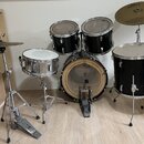 Sada akustických bubnů v černé barvě s kovovými obručmi. Obsahuje basový buben, dva tom-tomy, floor tom a stříbrný snare drum na stojanu. Na jednom z…