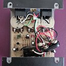 Fotografie zachycuje vnitřek elektronického zařízení v černém kovovém šasi. Uvnitř je viditelná zapojovací deska s komponenty a dvěma kulatými kovový…
