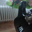 Fotografie zachycuje zadní část černého trupu elektrické kytary s viditelnou dřevěnou strukturou. V dolní části je otvor s vyvedenými strunami a v ho…