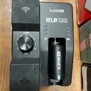 Černý bezdrátový systém Line 6 Relay G10S. Na zařízení je vidět nápis LINE 6 a model RELAY G10S, v držáku je umístěn vysílač s nápisem LINE 6. Vlevo…