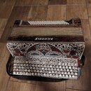 Akordeon s černým povrchem, zdobený bohatými červenými a bílými ornamentálními vzory. Na horní části je nápis KEDROLE zobrazený obráceně. Nástroj má…