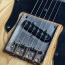 Detail elektrické kytary s přírodním dřevěným povrchem a černým pickguardem. Viditelný je chromovaný most s pružinami a jeden černý humbucker snímač.…