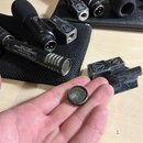 Na dlani je položen malý kulatý šedý mikrofonní kapsle s mřížkou. Vedle ní leží černý plastový držák s nápisy audio-technica a logem značky v kruhu.…