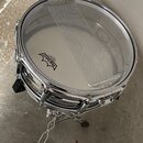 Stříbrný snare drum s kovovým obručováním a bílým membránou. Na membráně je černé logo REMO Ambassador Coated a nečitelný nápis Schmidtke. Přes membr…