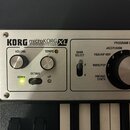Detailní pohled na ovládací panel černého syntetizátoru s nápisy KORG microKORG XL SYNTHESIZER/VOCODER. Panel je stříbrný a obsahuje knoflíky VOLUME…